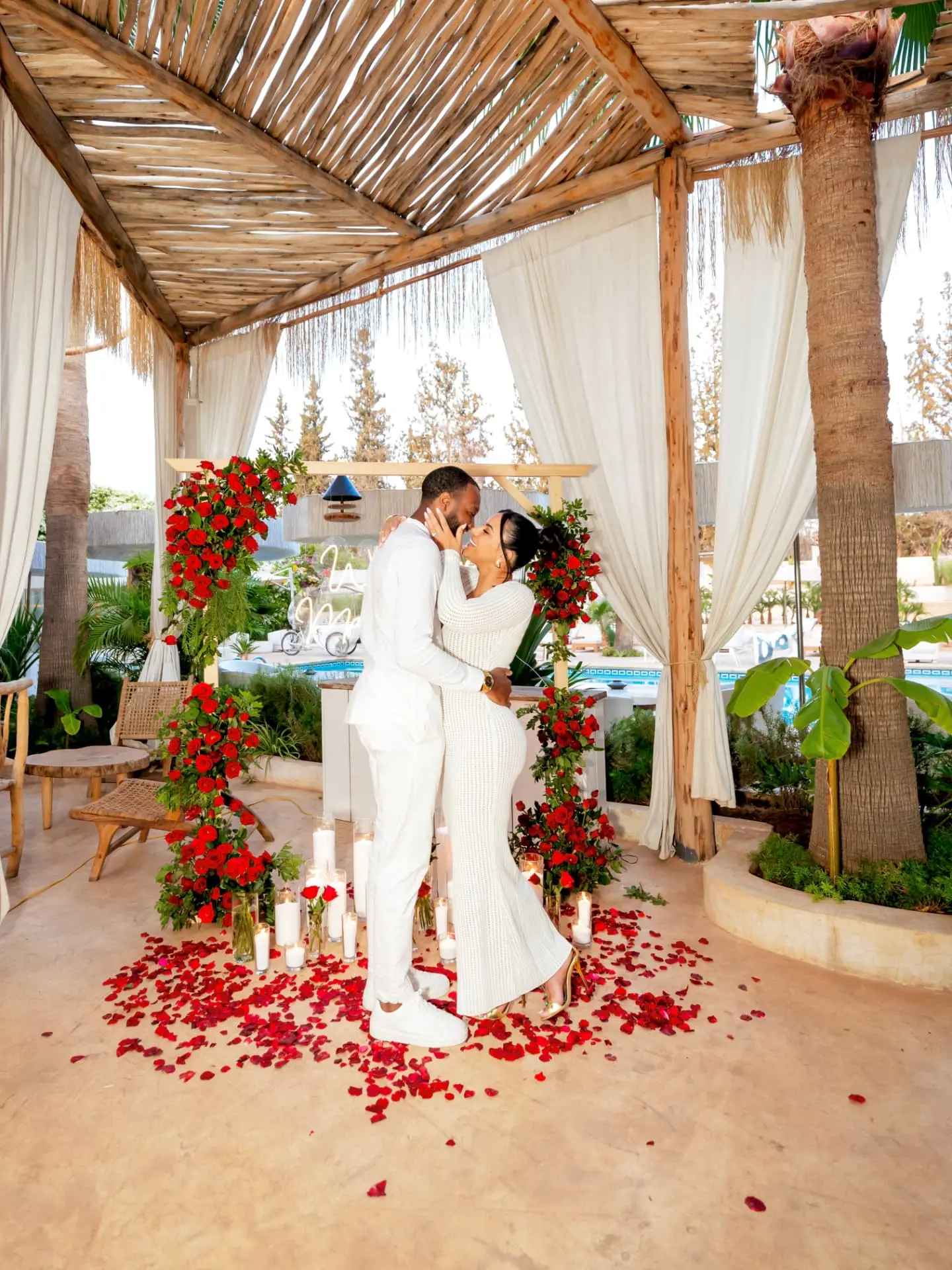 Kat and Elston embracing under a wooden pavilion draped with red roses and candles