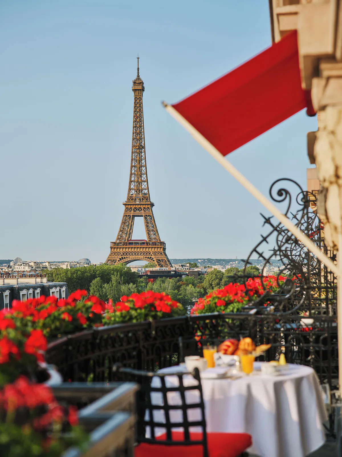 A sunlit Hôtel Plaza Athénée balcony framed by red geraniums and awnings, with the Eiffel Tower in the distance